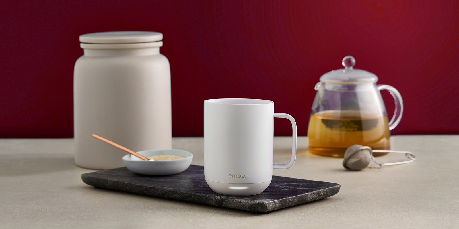 A white Ember Mug² sits on top of a dark marbled cutting board. There is a bowl of sugar to the left and a pot of tea to the right with a tea steeper. The background is dark red.