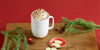 A white ember mug sitting on a wooden cutting board with a bowl of white chocolate disks, small pine cones, red ornaments, and festive branches on a red background.