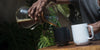 Person pouring Chemex brewed coffee into a black Ember Mug while it sits next to a white Ember Mug.