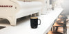 A latte filled Ember Mug² in black sits on a white coffee bar with a cream Strada espresso maker behind it