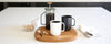 A black Ember Mug and white Ember Mug sit on a wooden cutting board next to a French Press filled with coffee. On the white countertop sits a small pitcher of cream and a bowl filed with brown sugar cubes.