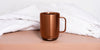 Ember Mug²: Copper Edition sitting on a light pink table with white blankets layered in the background.