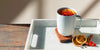 A white Ember Mug filled with deep orange tea sits on a white tray alongside candied orange slices, a cinnamon stick, a couple of cloves, and a handful of cranberries.