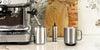 Stainless steel Ember Mug sits next to stainless steel milk frother, espresso machine, and cup, in front of wooden cutting boards on a cream marbled counter top.