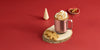 A Rose Gold Ember Mug² filled with whipped cream and cinnamon sits on a round cut of wood along wih two gingerbread cookies. The background is a deep red and is filled with gingerbread trees and cinnamon sticks.