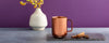 Ember Mug: Copper Edition on a dar gray counter with a cream vase and purple background.